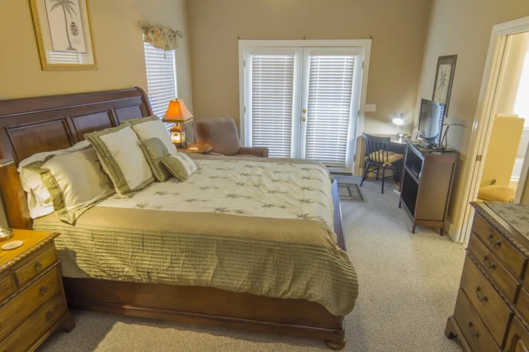 King-size wooden sleigh bed with palm-patterned bedding next to beige armchair and French doors with white blinds.