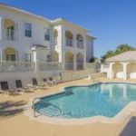 Curved swimming pool with light blue tile border, surrounded by tan deck and black lounge chairs.