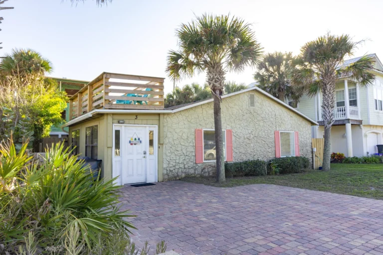 Paved driveway leads to the one-level house with textured beige walls, coral window shutters, and a rooftop deck with wooden railing.