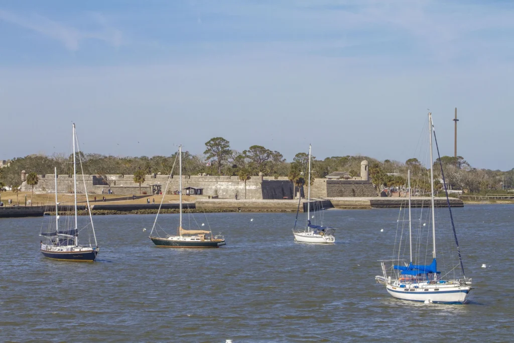 Four sailboats float on calm water with a historic stone fort and palm trees lining the shore beyond.