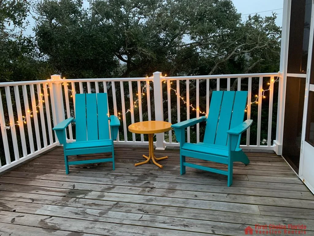 Two turquoise Adirondack chairs flank a round yellow metal table on the screened balcony with string lights.