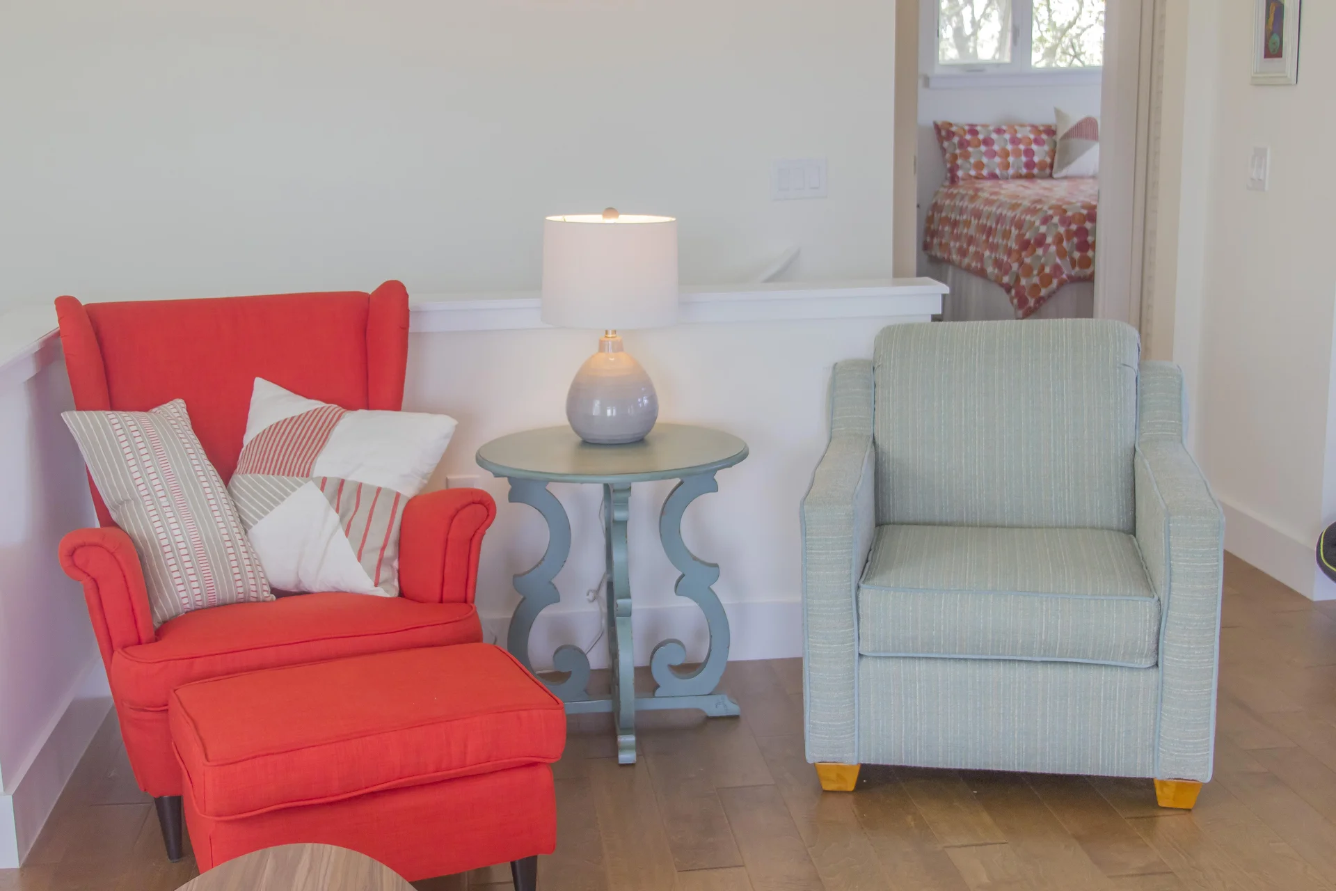 Seating area with a red wingback chair, matching ottoman, light blue armchair, and round side table with lamp.
