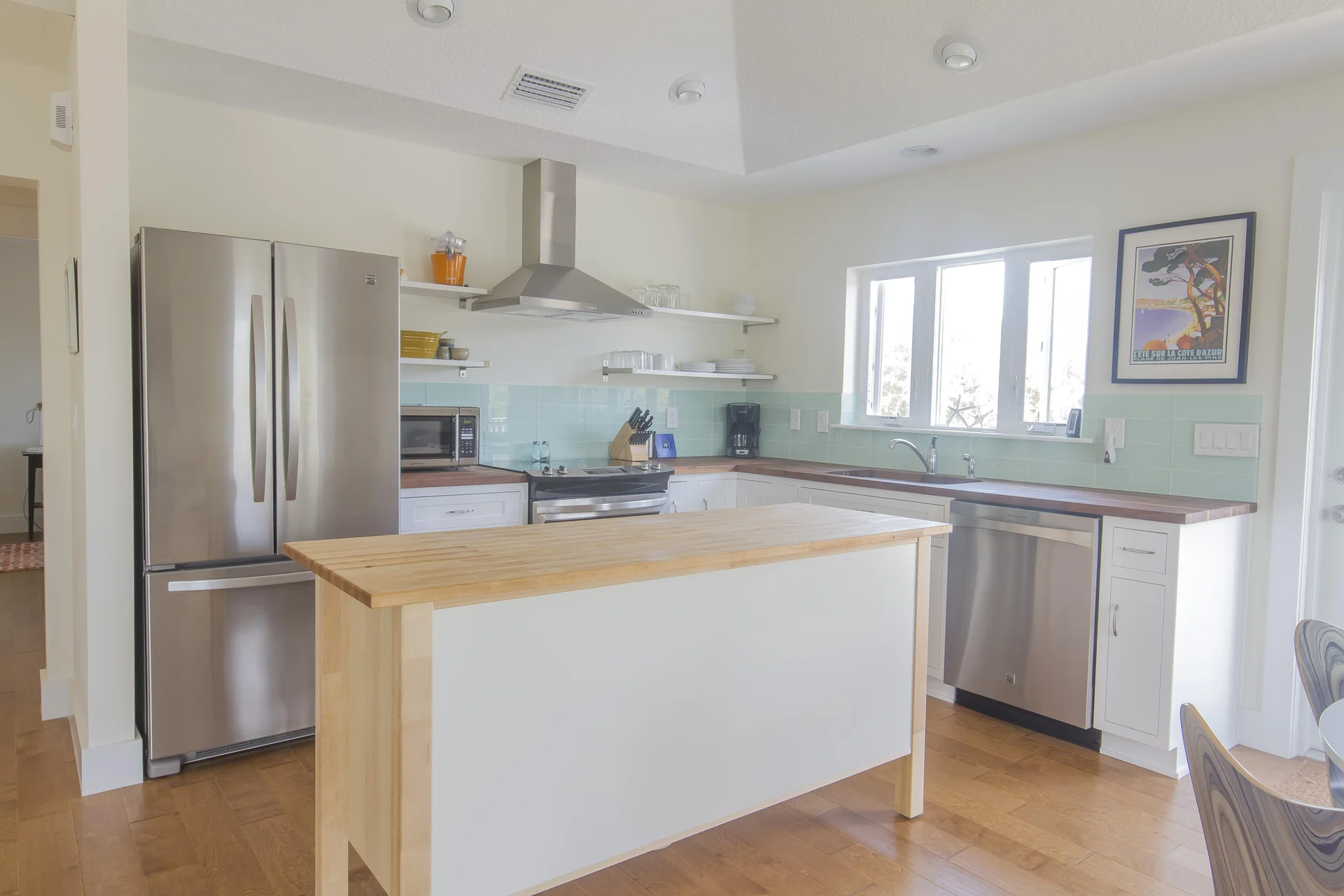 Butcher block island stands center in the kitchen with stainless steel appliances and pale aqua tile backsplash.