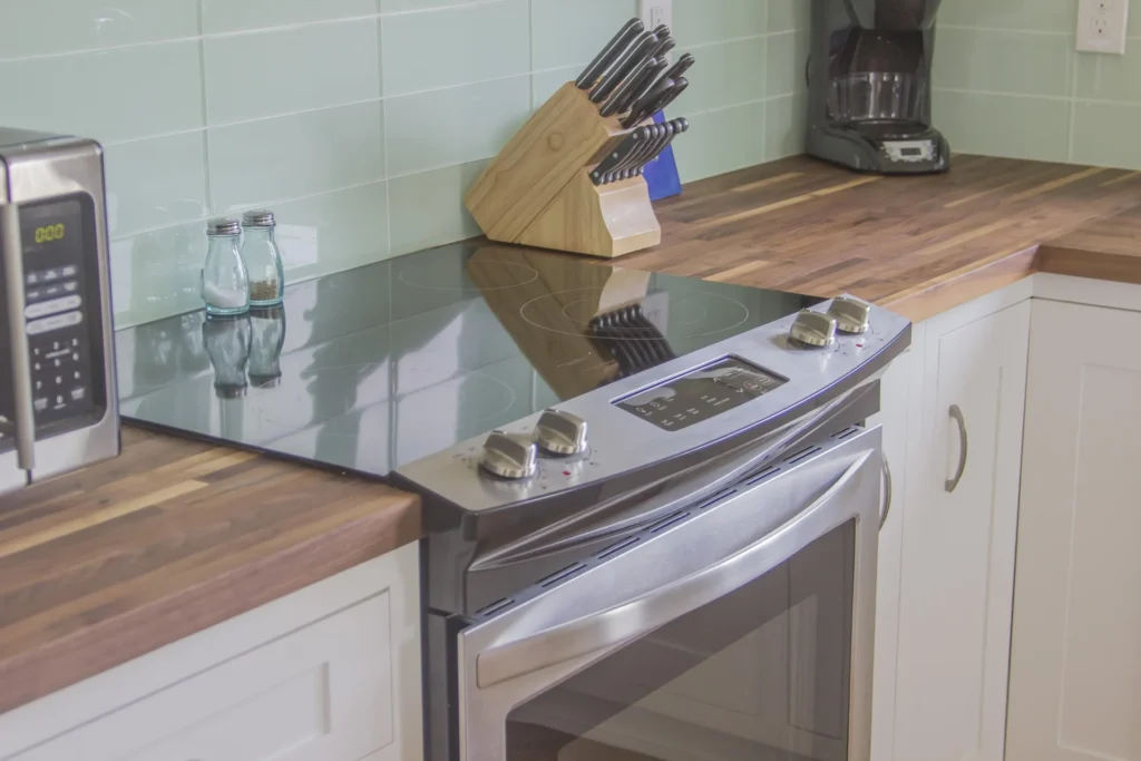 Stainless steel electric cooktop and oven set into white cabinetry with butcher block countertops and pale green tile backsplash.