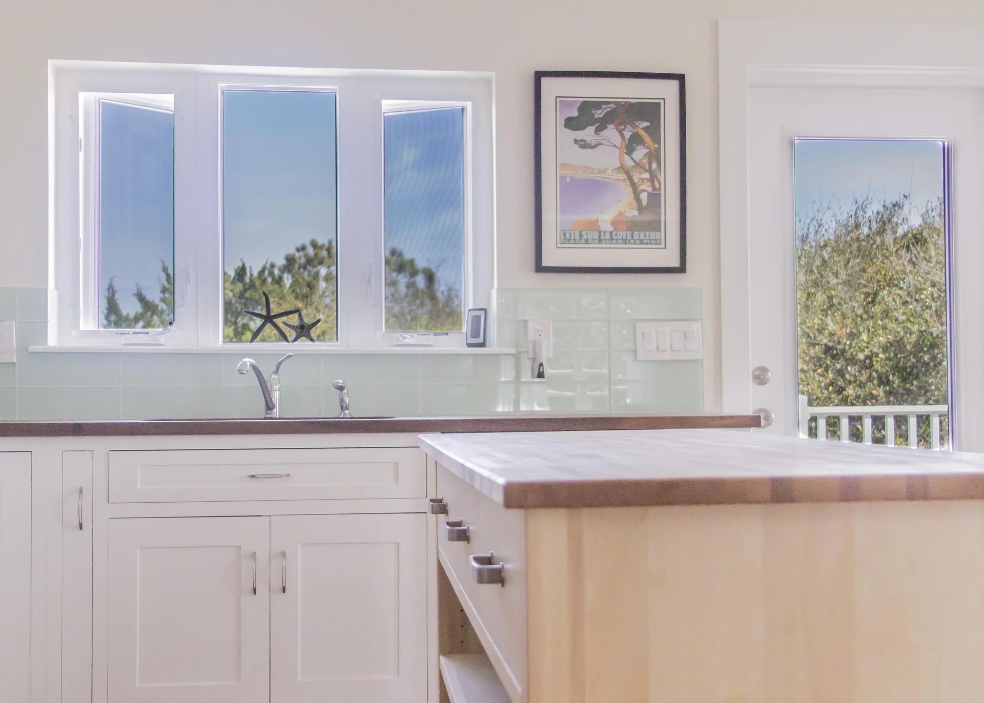 Natural light filters through three white-framed windows above a double sink with a wooden countertop and pale green tile backsplash.