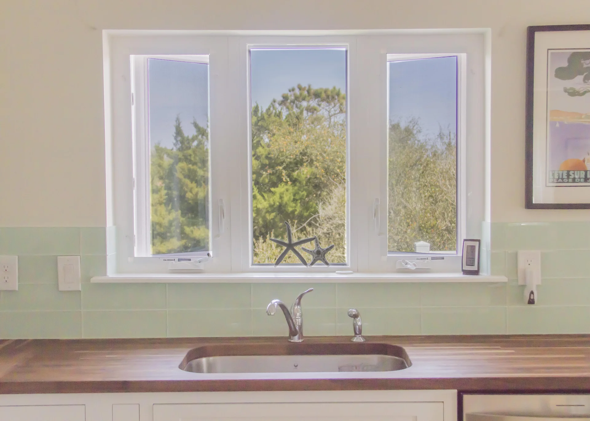 Kitchen sink with polished chrome faucet beneath triple-pane windows overlooking green trees and starfish decor.