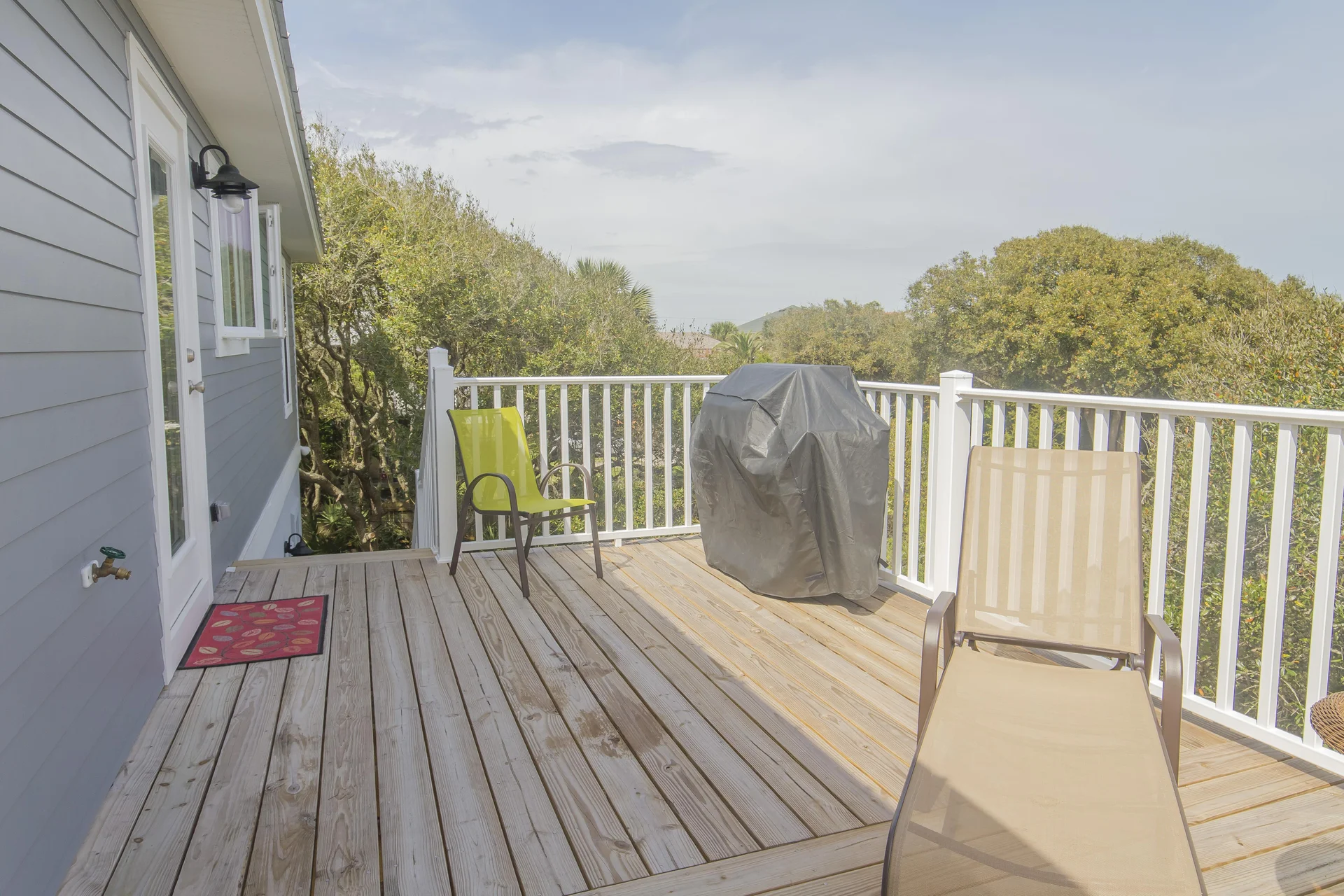 Second-floor deck with natural wood planks, white railing, two outdoor chairs, and covered grill overlooking trees.