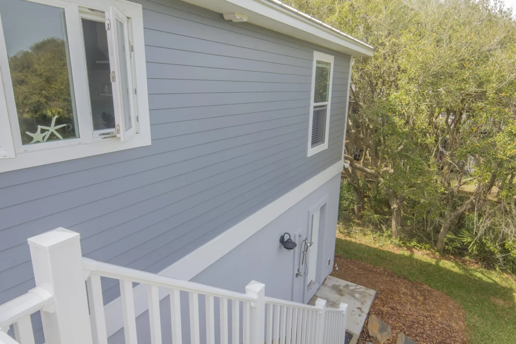 Exterior side view of the house with gray siding, white trim, and a white railing overlooking a wooded backyard.