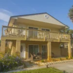 Two-story villa exterior with upper deck featuring beige railings and patio seating below on textured stucco pillars.