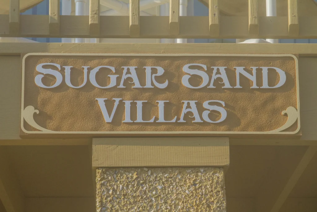 Close-up of textured stucco sign with white raised letters spelling "Sugar Sand Villas" mounted on beige wooden beam.