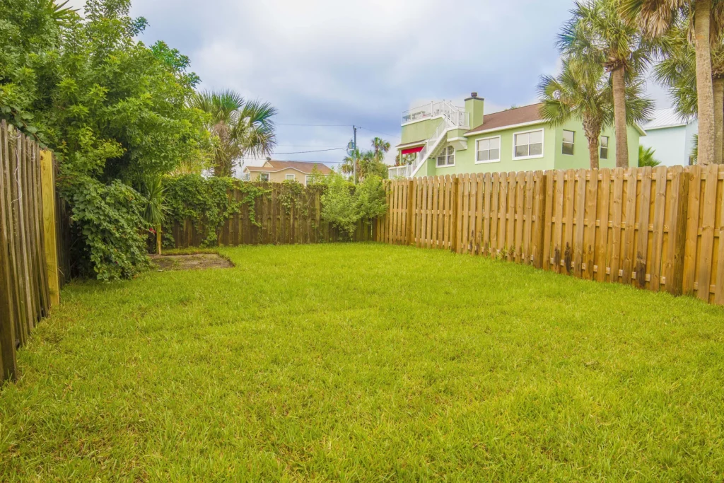 Fenced backyard with green lawn bordered by wooden planks and leafy shrubs under a cloudy sky.
