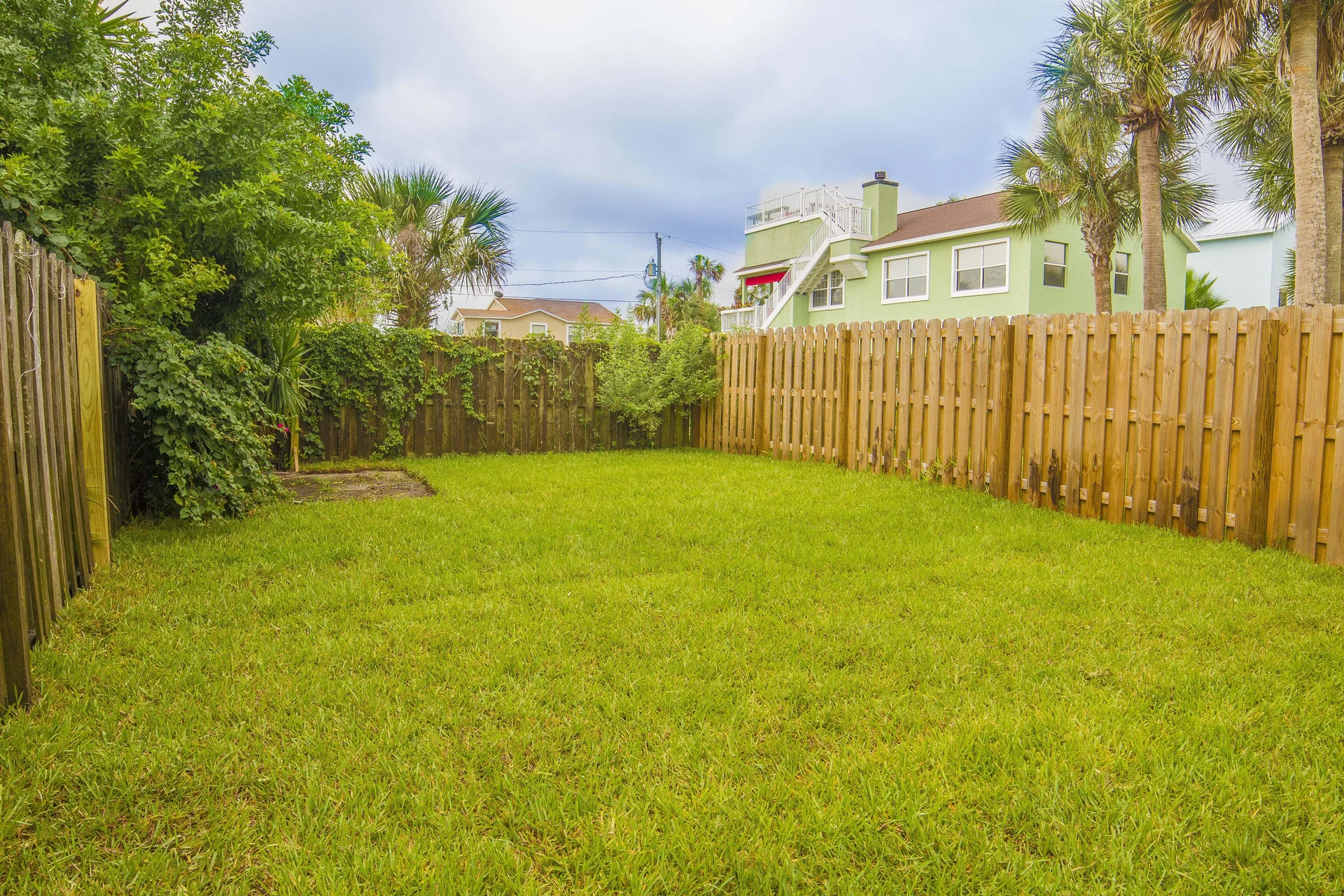 Fenced backyard with green lawn bordered by wooden planks and leafy shrubs under a cloudy sky.