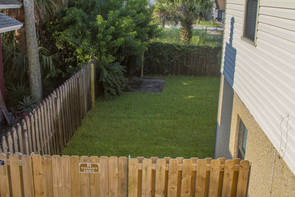 Fenced backyard with grass lawn bordered by wooden picket fences and dense greenery along one side.