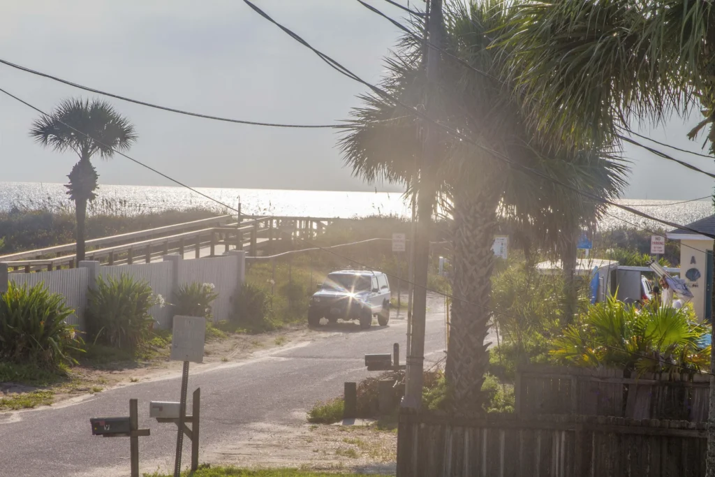 Sunlight reflects off the ocean beyond a paved road lined with palm trees, wooden mailboxes, and sandy beach access.