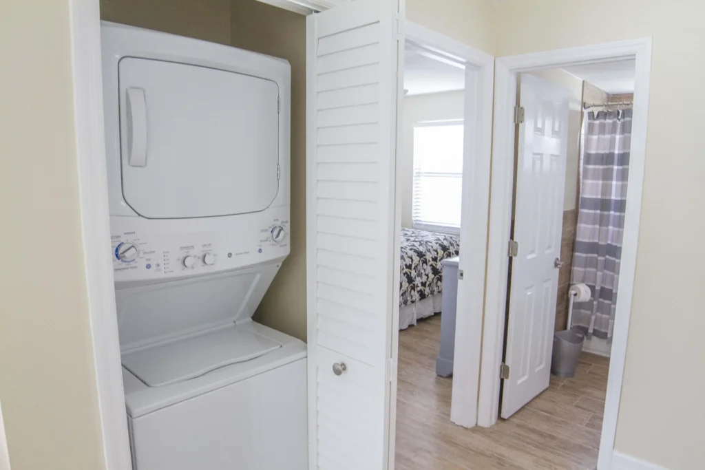 Stacked white washer and dryer fit into a closet with louvered doors beside the hallway leading to bedroom and bathroom.