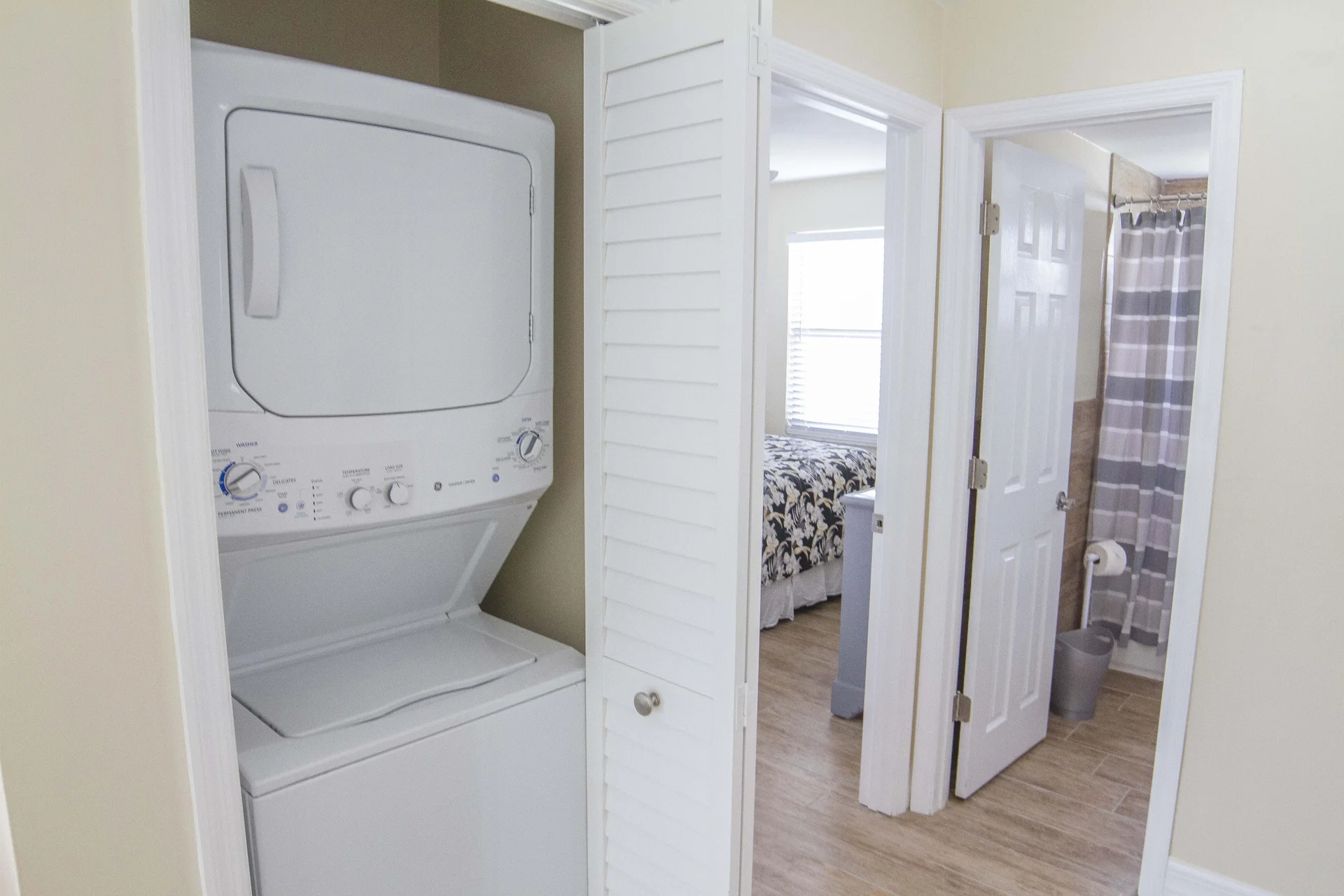 Stacked white washer and dryer fit into a closet with louvered doors beside the hallway leading to bedroom and bathroom.