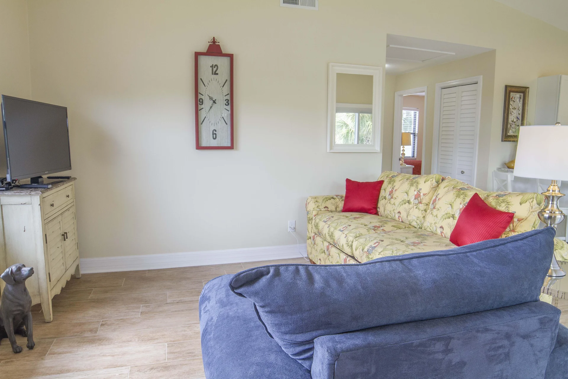 Blue armchair and floral sofa with red pillows face a flat-screen TV on a distressed white wooden cabinet.