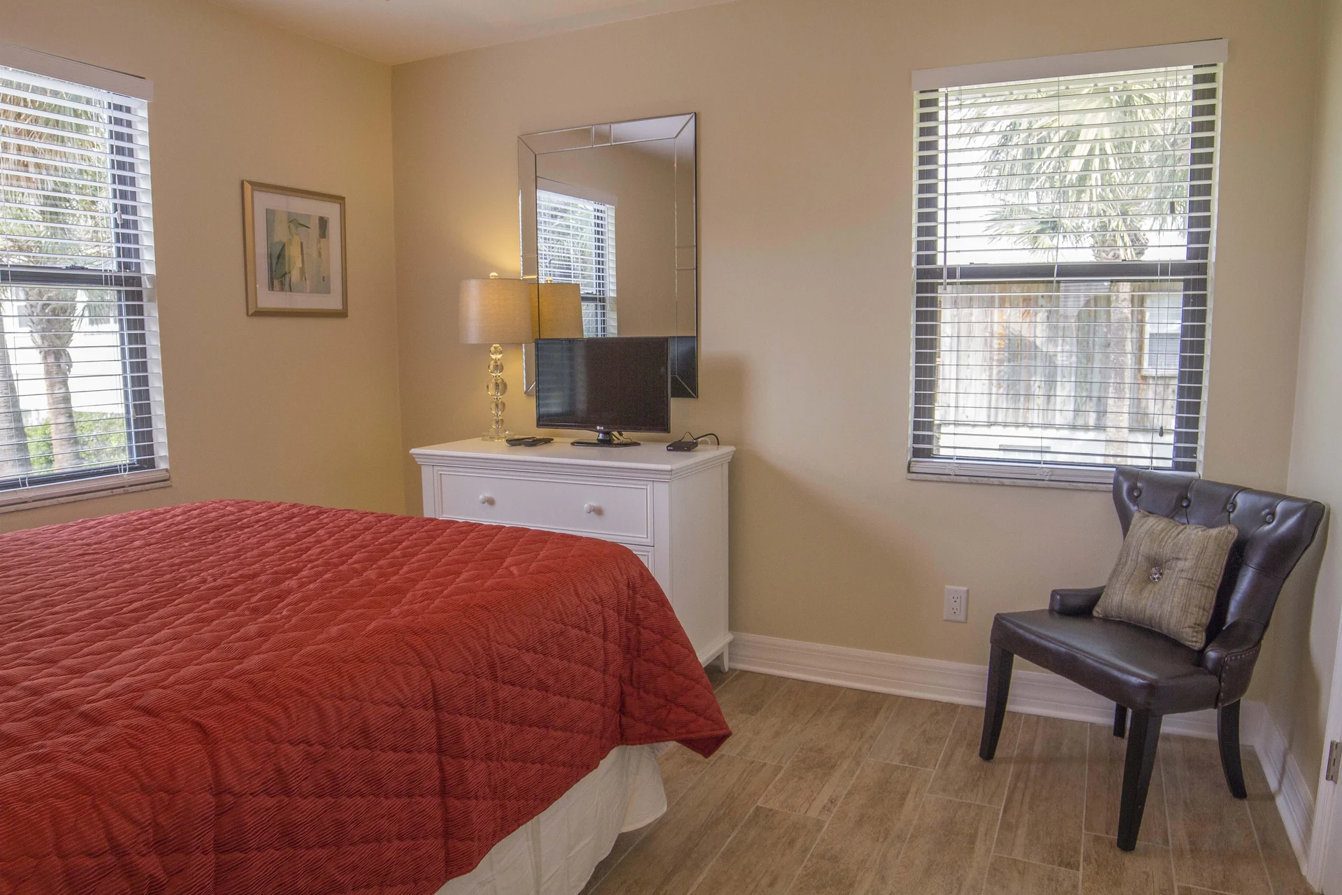 Bedroom corner with red quilted bedspread, white dresser holding a lamp and TV, and a black leather accent chair.