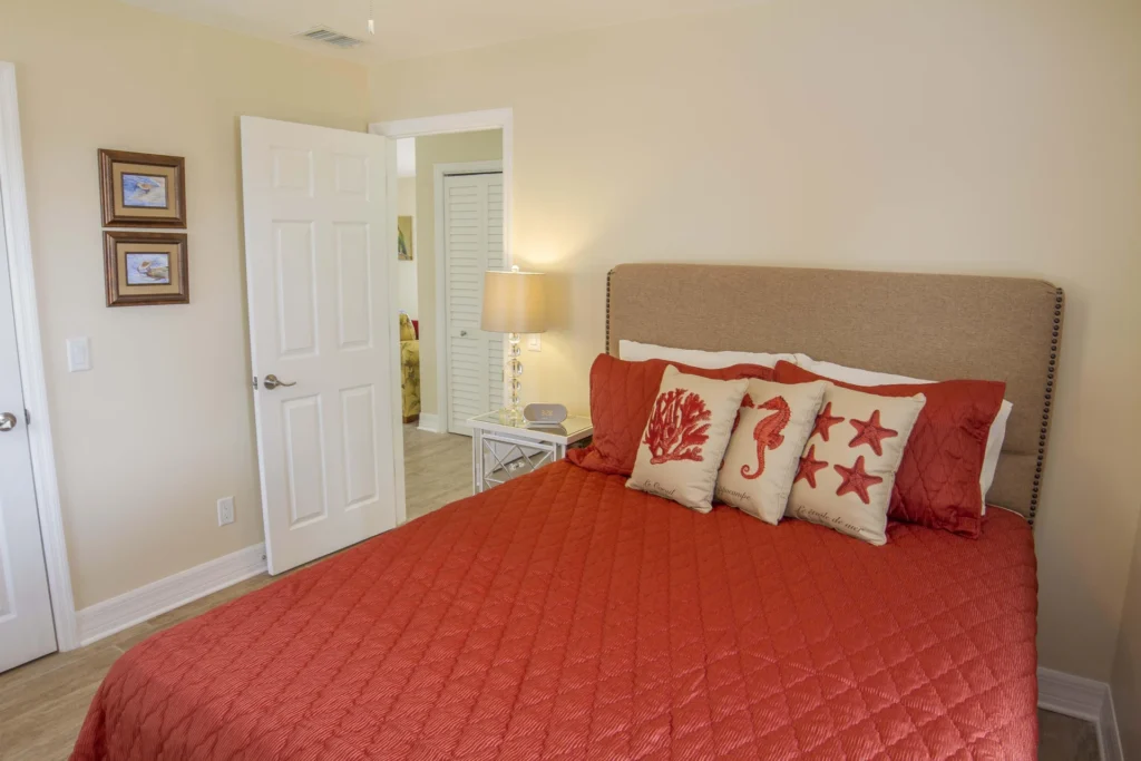 Bedroom with beige upholstered headboard, coral quilted bedding, sea-themed pillows, and a glass table lamp.