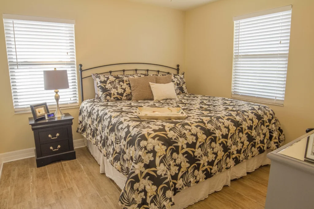 King bed with floral comforter sits between two windows, flanked by a black nightstand and silver lamp.