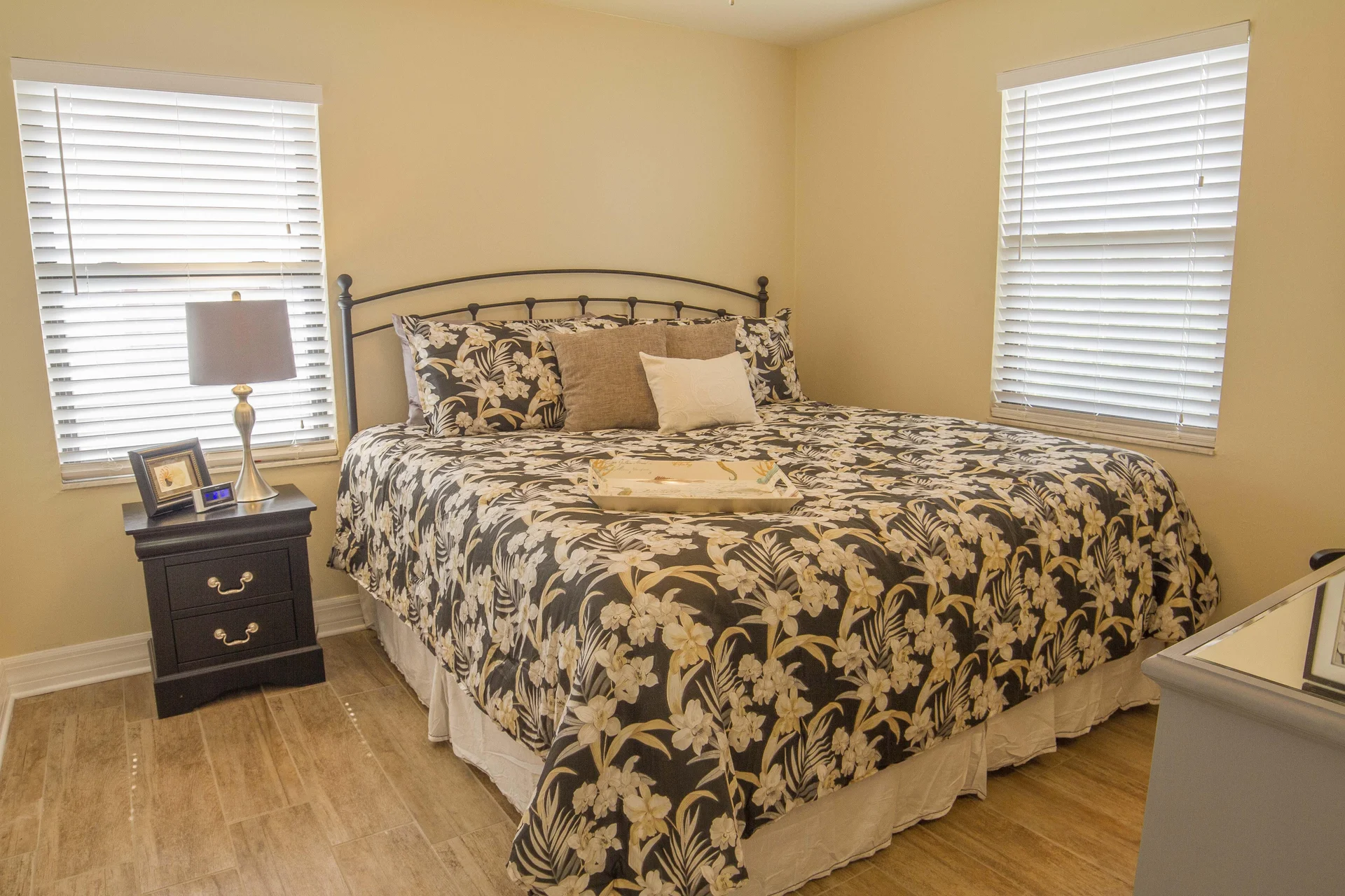 King bed with floral comforter sits between two windows, flanked by a black nightstand and silver lamp.