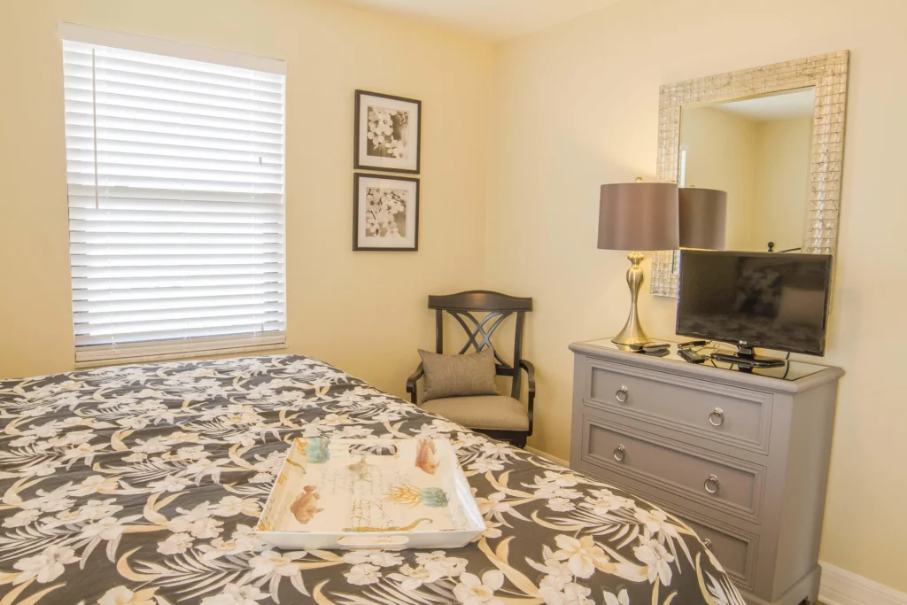Floral-patterned bedspread covers bed beside black wooden chair and gray dresser with lamp and TV.
