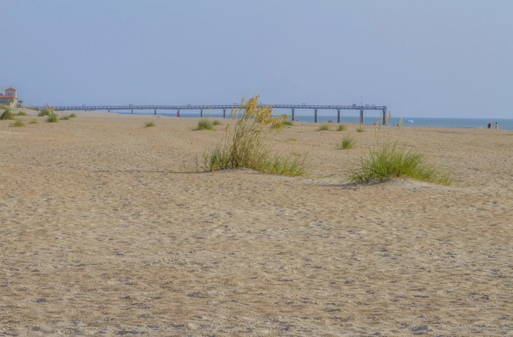 Sandy beach stretches toward a wooden pier with sparse sea oats and calm ocean waters under a clear sky.