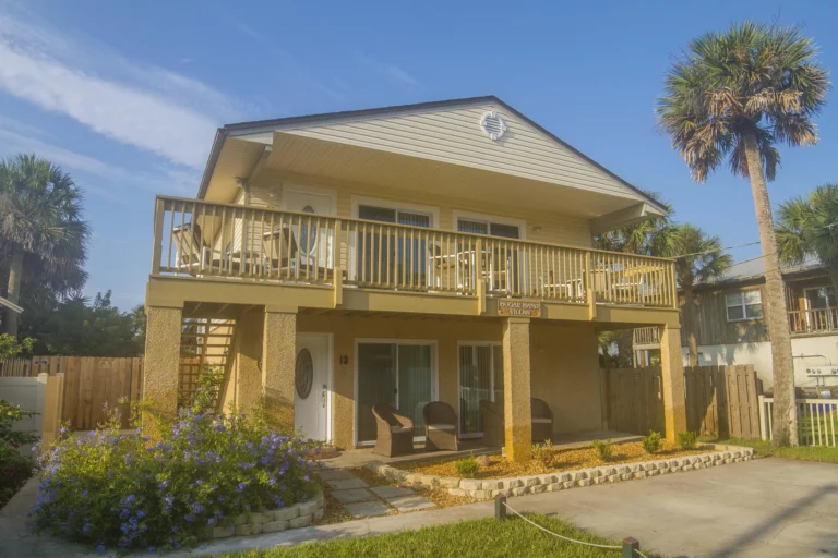 Ground floor patio shaded by the upper deck, furnished with wicker chairs and surrounded by flower beds.