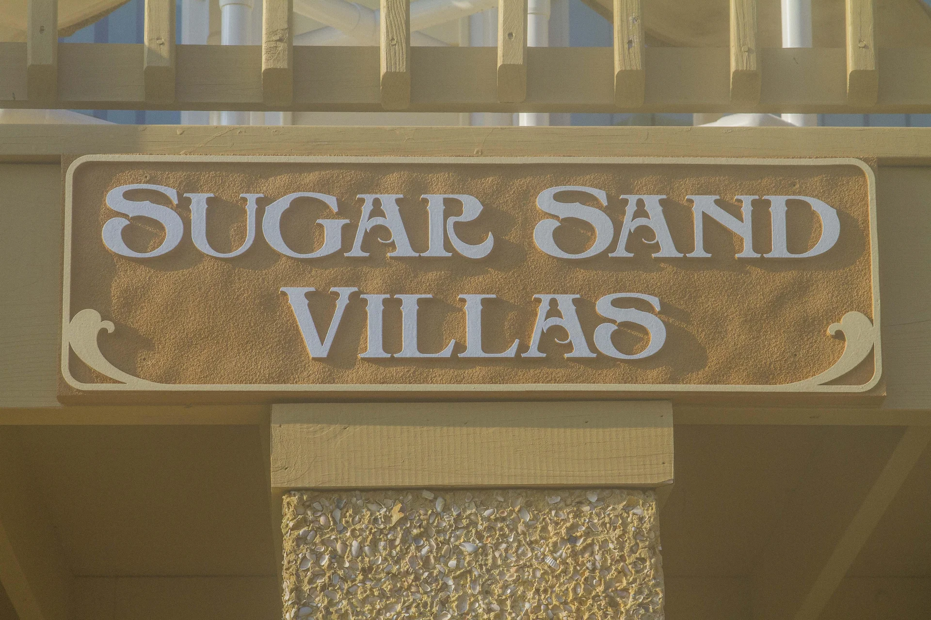Close-up of textured tan sign with raised white letters reading "Sugar Sand Villas" mounted on wood and shell-embedded concrete.