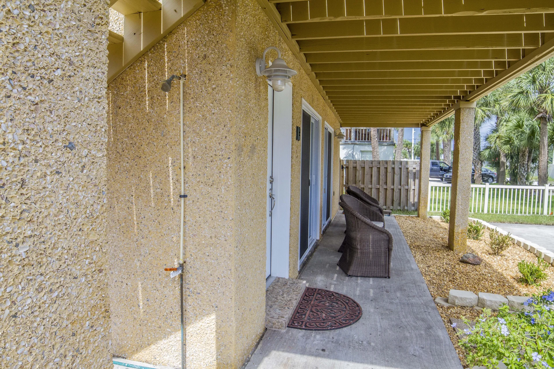 Concrete covered patio with two dark wicker chairs, textured shell walls, outdoor shower, and garden beds.