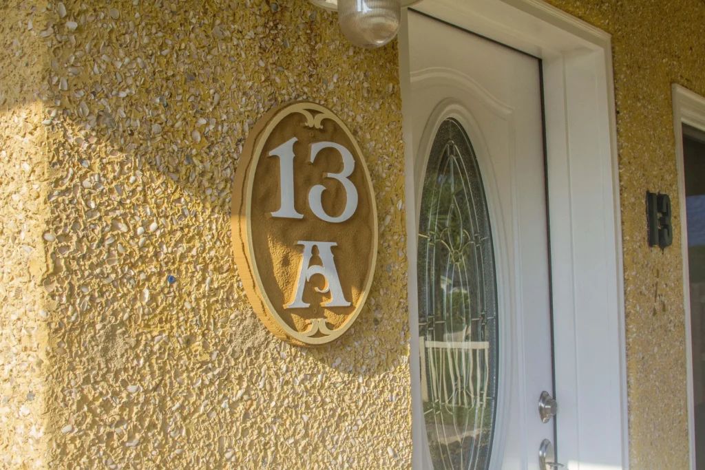 Yellow pebble-textured exterior wall with oval house number plaque beside white front door with decorative glass panel.