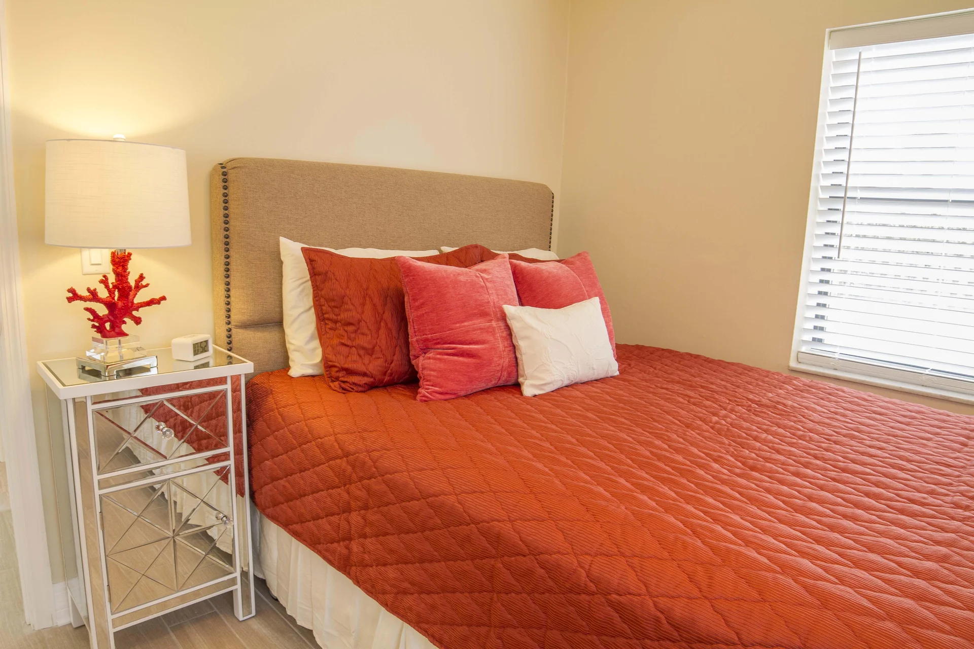 Bed with a beige upholstered headboard, rust-colored quilt, pink and white pillows, and mirrored nightstand with coral lamp.