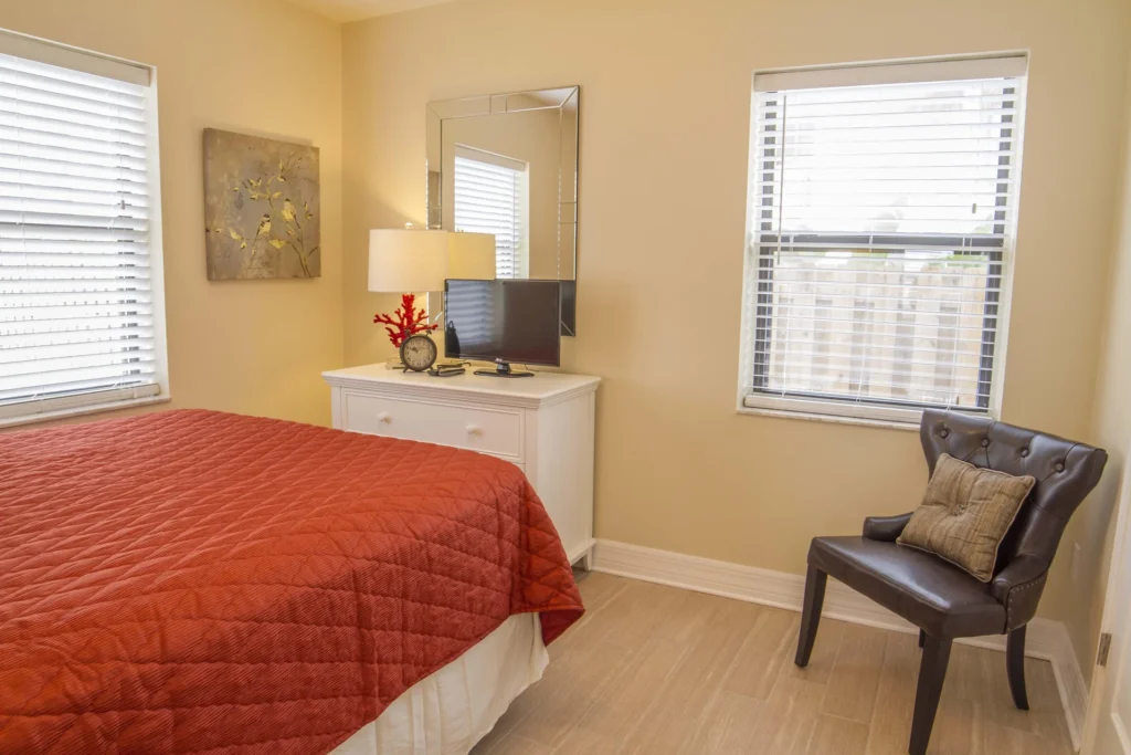 Bedroom with warm beige walls, two windows with blinds, a white dresser holding a TV and lamp, and a dark leather chair.