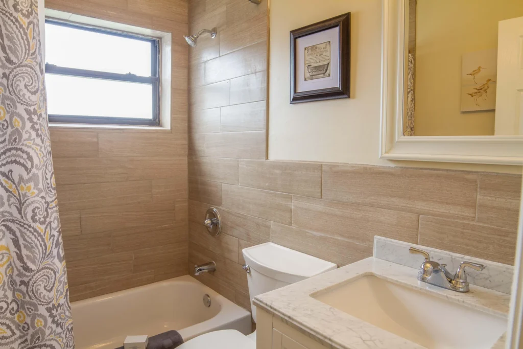 Rectangular beige tiles surround the tub and shower with a patterned fabric curtain and frosted window.