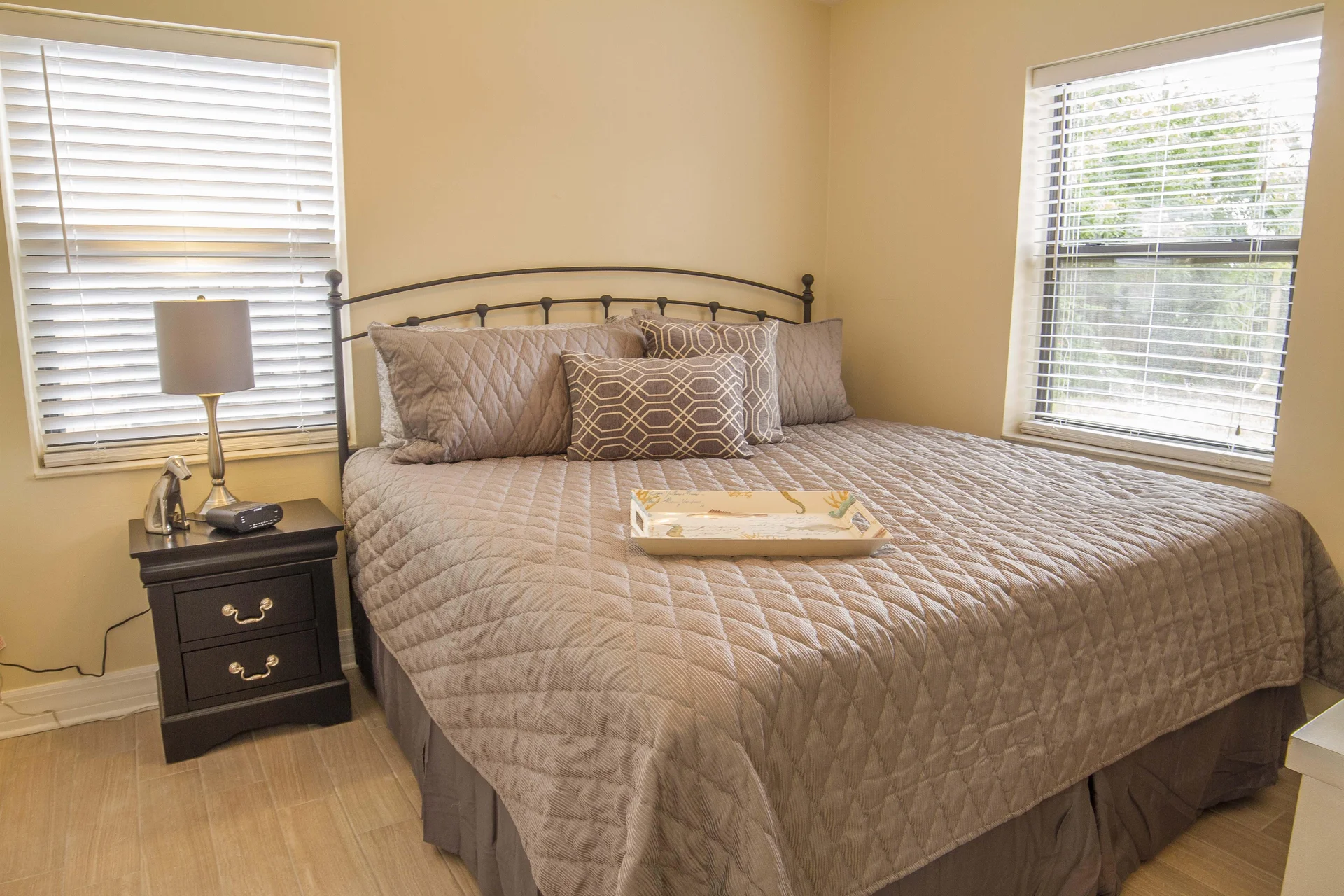 King bed with quilted taupe bedding and patterned pillows between two windows with white blinds.