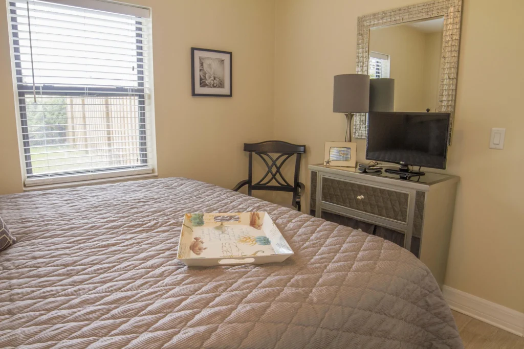 Beige quilt covers the bed with a decorative tray, beside a black wooden chair and mirrored dresser with TV.