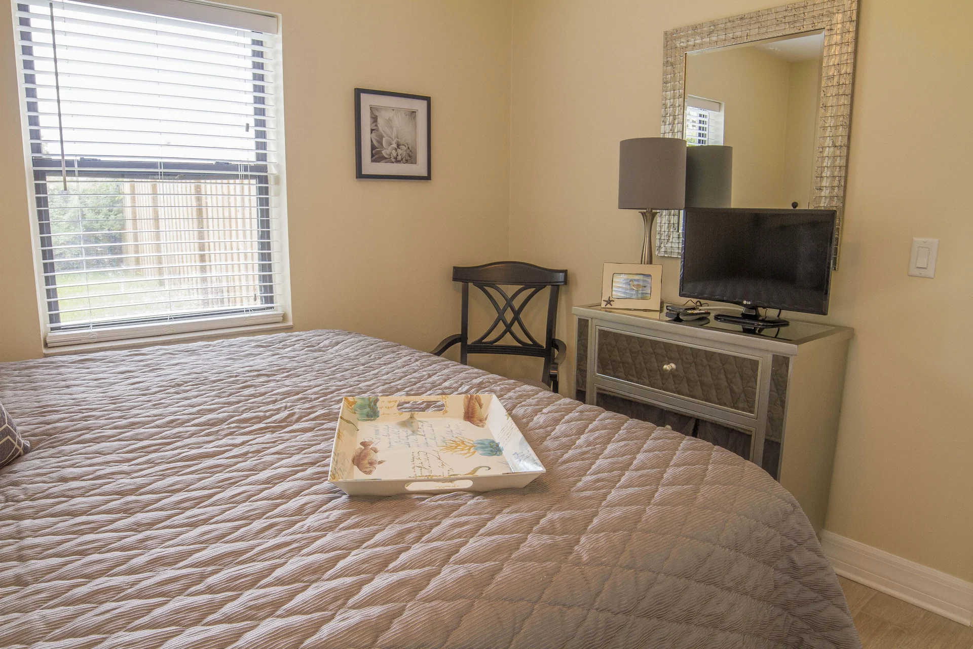 Beige quilt covers the bed with a decorative tray, beside a black wooden chair and mirrored dresser with TV.