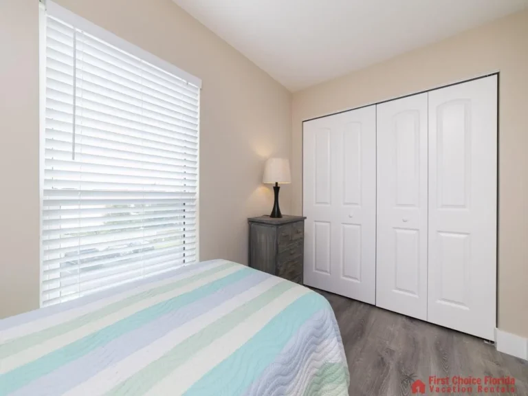 Natural light filters through white blinds onto the striped pastel quilt and wood-look vinyl flooring near the closet.