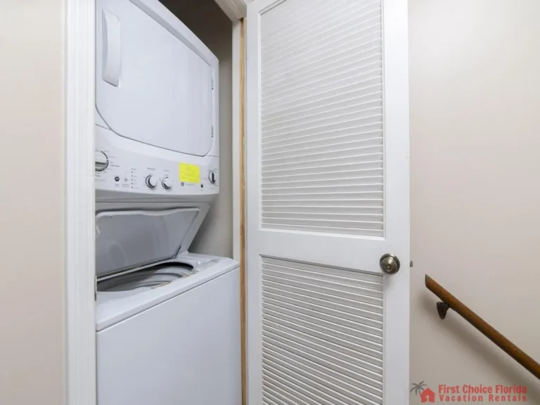 Stacked washer and dryer tucked into a narrow closet with white louvered door and beige walls.