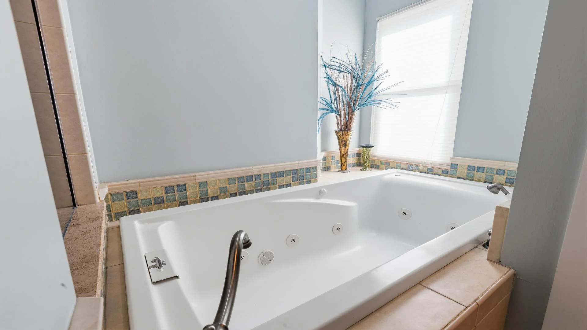 A white jetted bathtub features chrome fixtures and a tiled ledge with blue and green mosaic accents.
