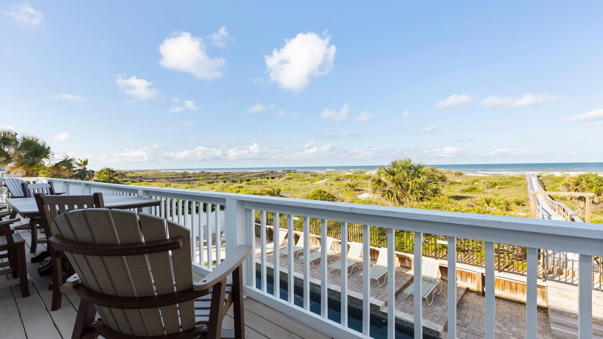 Wood deck balcony with Adirondack chairs and high tables overlooks pool, dunes, and ocean beyond.