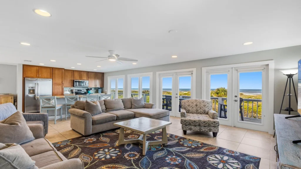 Oceanfront living area with sectional sofa, patterned armchair, floral rug, and kitchen with wood cabinets.