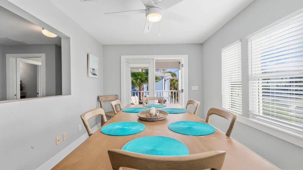 Dining area with natural wood table, turquoise placemats, light wood chairs, and white blinds over windows.
