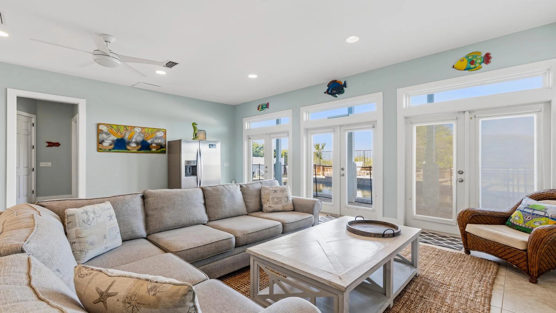 Light blue walls frame a sectional sofa and wicker chair facing a white wooden coffee table near glass doors with ocean views.