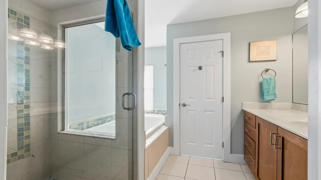 Glass-enclosed shower features beige tiles with blue mosaic accents beside a soaking tub and wood vanity.