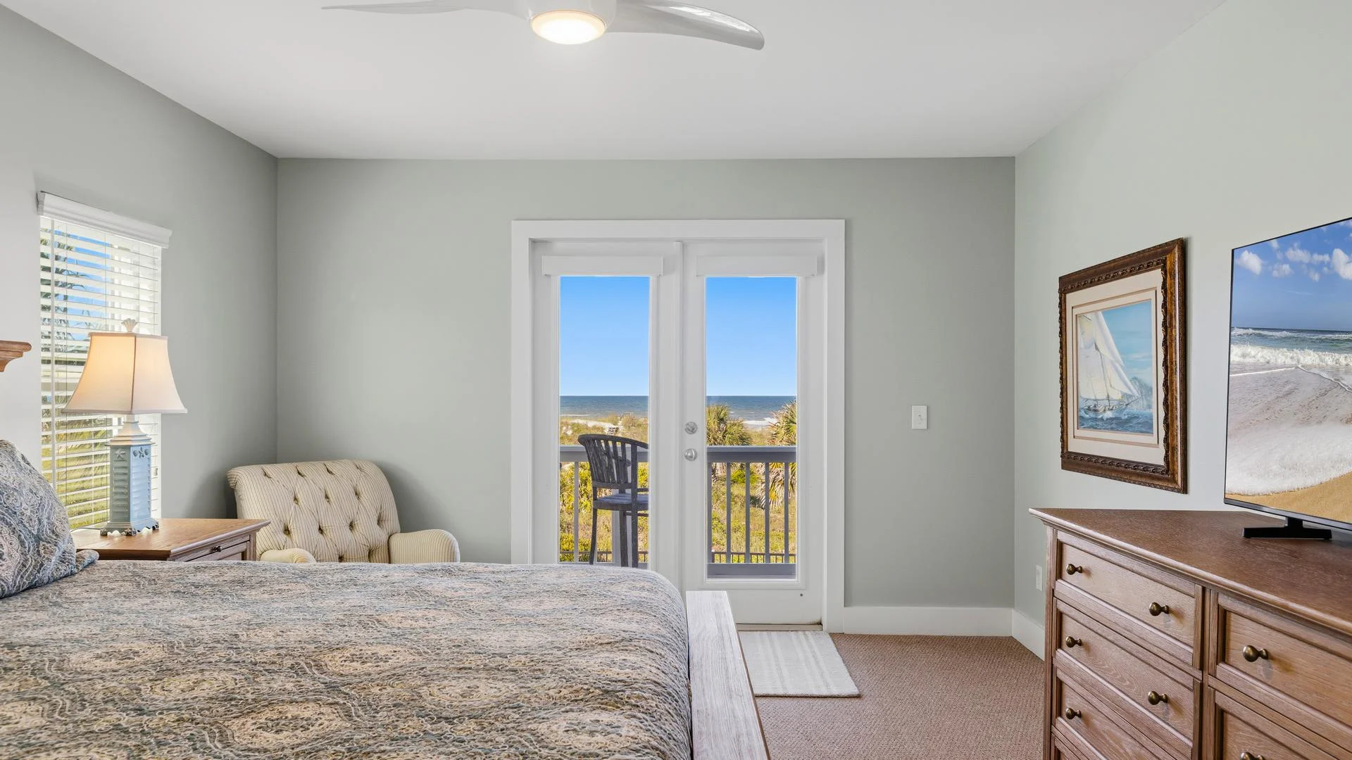 Bedroom with light gray walls, wooden dresser, patterned bedding, and glass doors opening to an ocean view balcony.