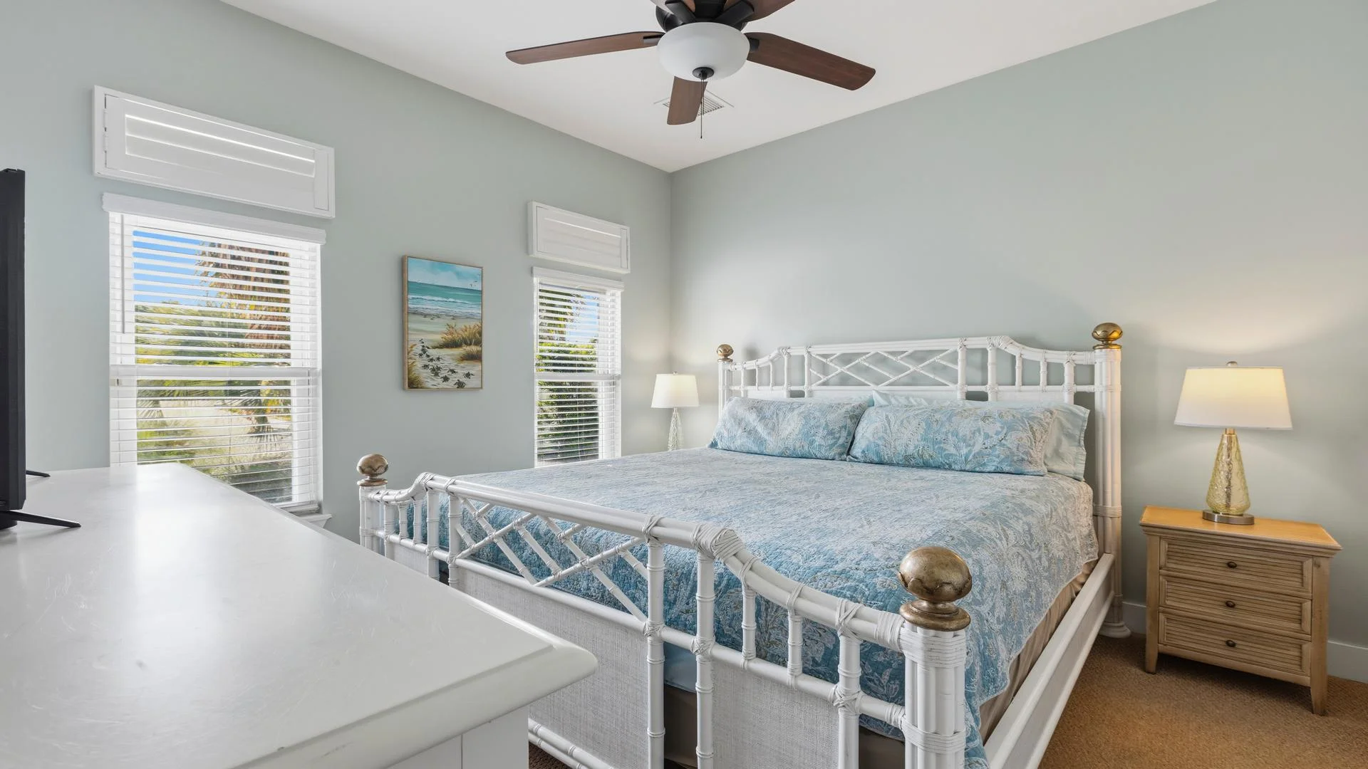 First-floor light-filled bedroom with white rattan bedframe, blue patterned bedding, wooden nightstand, and ceiling fan.