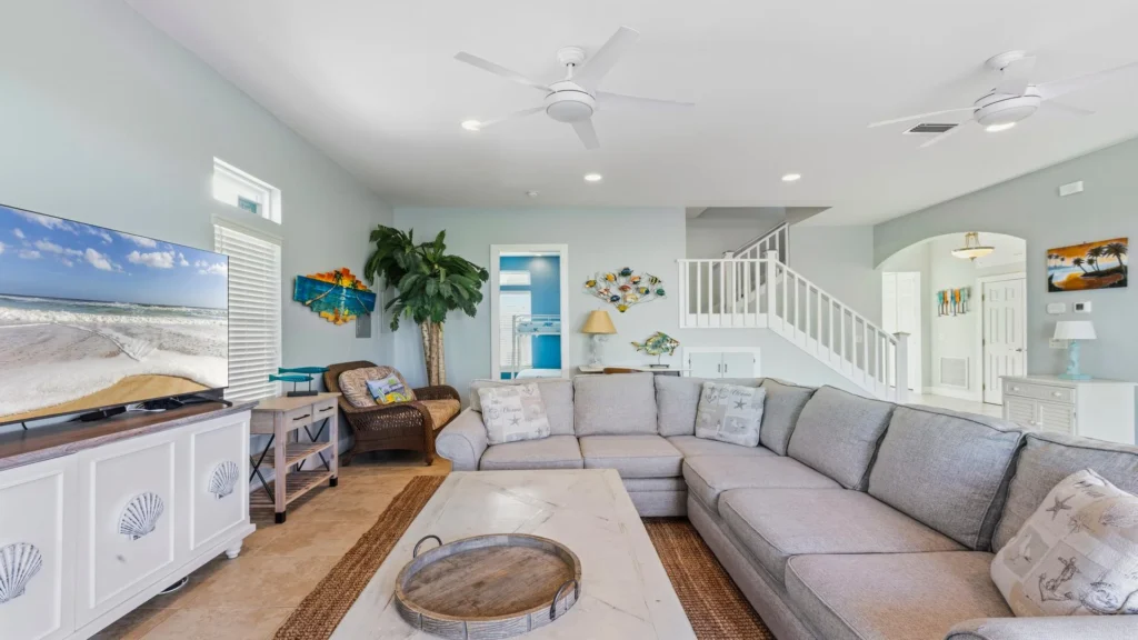 Living room with a large gray sectional sofa, white shell-accented TV console, and tropical wall art near staircase.