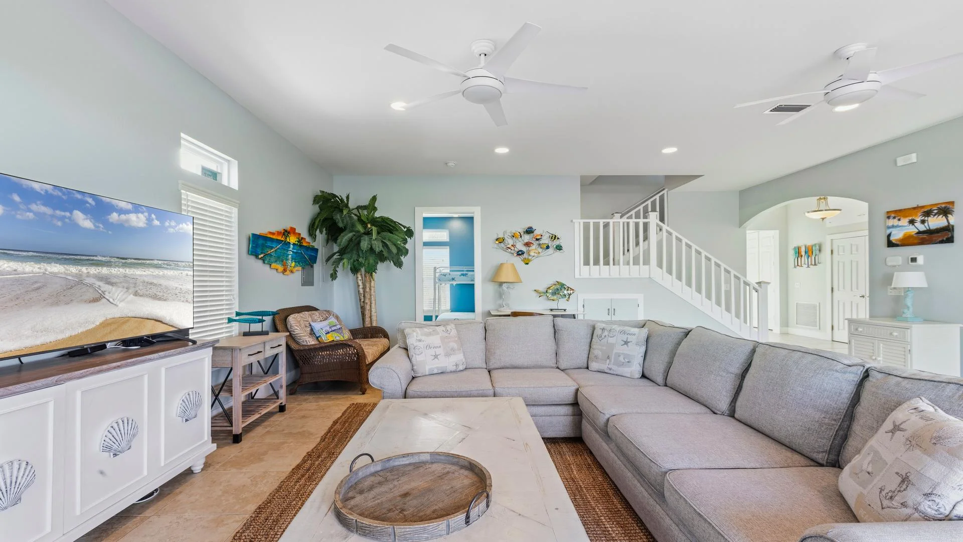 Living room with a large gray sectional sofa, white shell-accented TV console, and tropical wall art near staircase.