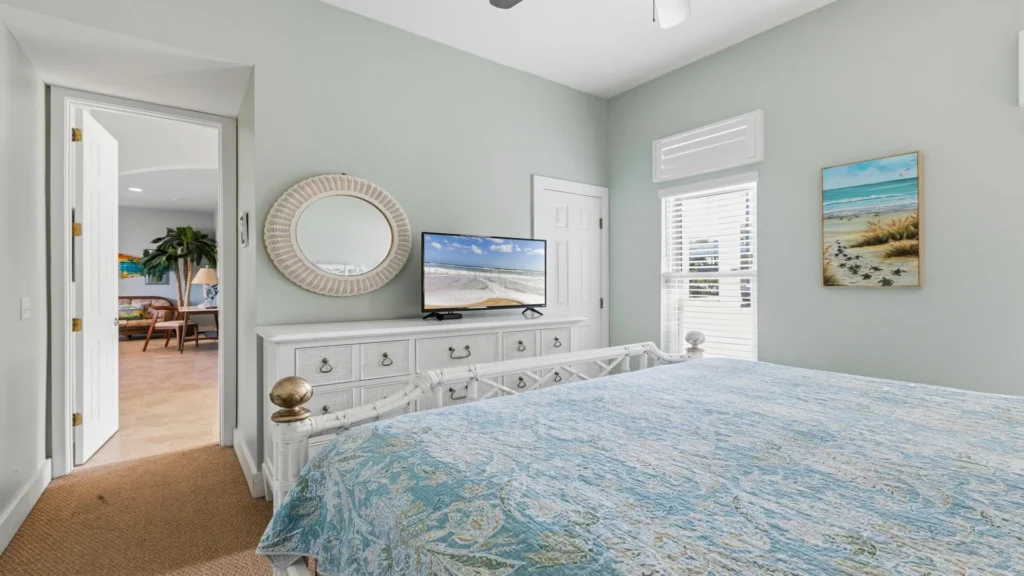 Bedroom with white rattan bed frame, blue floral quilt, dresser topped by round wicker mirror and flat-screen TV.